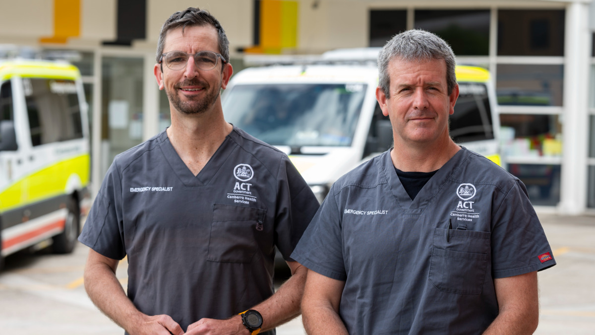 Two males doctors stand near the ambulance bay outside Canberra Hospital Emergency Department. They are wearing grey scrubs. 