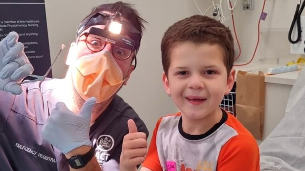 A doctor wearing gloves, a headlamp, face shield and mask holds up a small piece of Lego with tweezers. He is kneeling next to a small child. Both are giving thumbs up.
