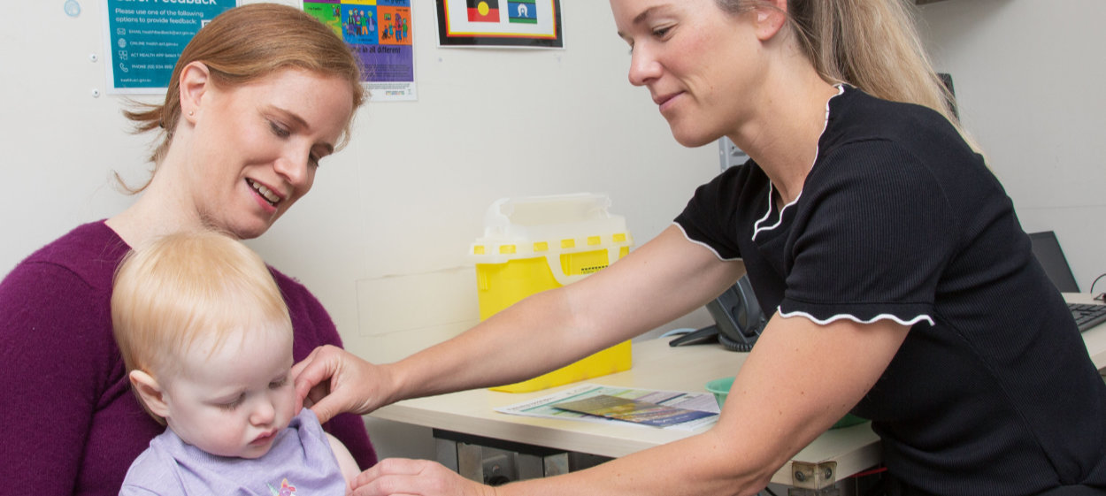 A nurse prepares to administer a vaccination to a young toddler sitting on their parent's lap in a medical clinic.