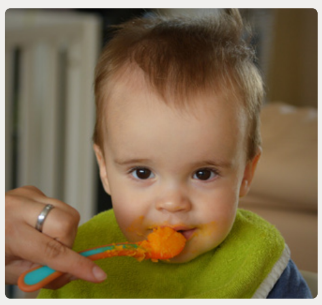 A young child with short hair wearing a green bib is being fed orange food with a spoon by a adult
