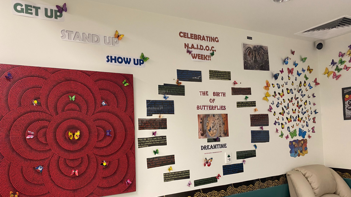 Two walls are decorated to celebrate NAIDOC Week. There is a sign saying 'the Birth of Butterflies' surrounded by text with a Dreamtime story. There are many colourful, paper butterflies of different sizes across the walls and on a nearby Aboriginal artwork. 