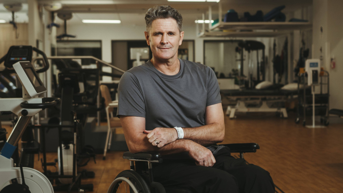 Chris sits in a wheelchair in a rehabilitation gym. He is smiling at the camera.
