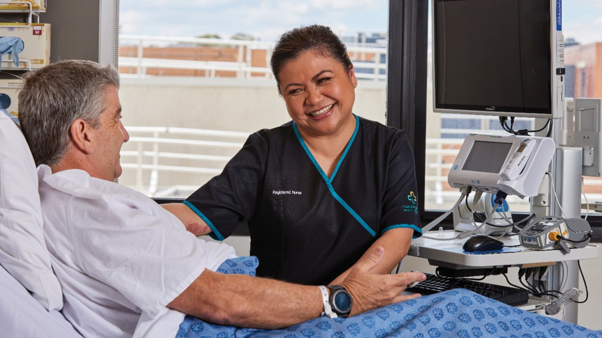 A patient is in bed at Canberra Hospital. He is wearing a hospital gown and looking at a nurse, who is smiling at him.