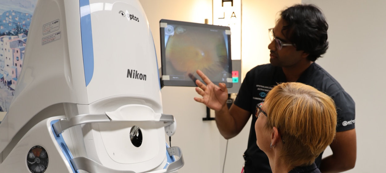 A clinician explains a wide-field retinal scan displayed on a monitor to a patient, with the Optos Silverstone RGB ultra-wide field imaging device in the foreground at Canberra Hospital Eye Clinic.