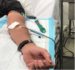 Patient's forearm resting on a hospital bed with an intravenous (IV) cannula inserted and secured with dressing, connected to IV tubing. A green tourniquet is visible above the insertion site, and medical equipment is positioned nearby.