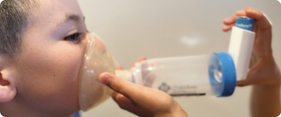 A child is pictured with an asthma puffer and spacer covering their mouth and nose. An adult is standing out of view to help the child ensure there are no gaps around the edges of the mask.