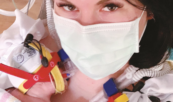 Close-up of a woman wearing a surgical mask, with her newborn baby in an upright position on her bare chest in a hospital setting.