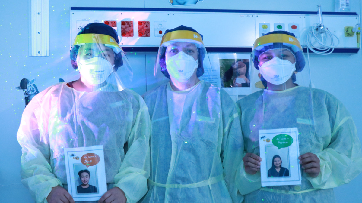 Three COVID-19 nurses look at the camera while wearing PPE. They are holding signs with their photos and names.