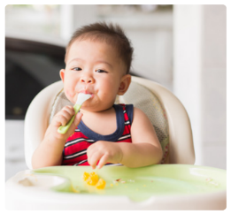 A young child in a red and navy blue striped sleeveless shirt sits in a high chair, holding a green spoon. Some food is visible on the green tray in front of them, behind them is a blurred indoor background.