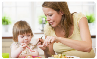An mother and a child sit at a table in a bright room with windows and green plants. The mother is holding a knife and fork, assisting the child with food on the table.