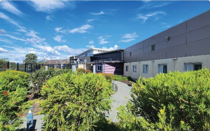 Modern Dhulwa Mental Health Unit building surrounded by landscaped gardens, with a pathway leading to the entrance under a blue sky