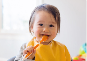 A young child wearing a yellow bib is eating with an orange spoon. Some food is visible on the child's face. The background is blurred, indicating an indoor setting with natural light from a window.