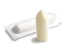 A white, bullet-shaped suppository standing upright in front of a partially opened silver-and-white blister package on a light background. The suppository has a smooth, rounded tip and a matte finish