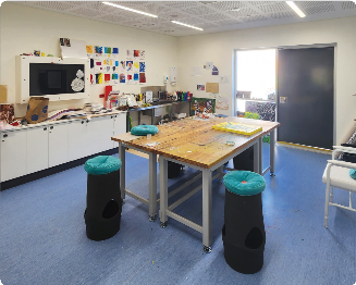 A small learning and group therapy area. A medium sized, square table sits in the middle of the area, with tall, cushioned stools surrounding the table. A wet area with a sink is located on a back wall, painted artworks are on the walls above it. A tv and cupboard area is next to the wet area.