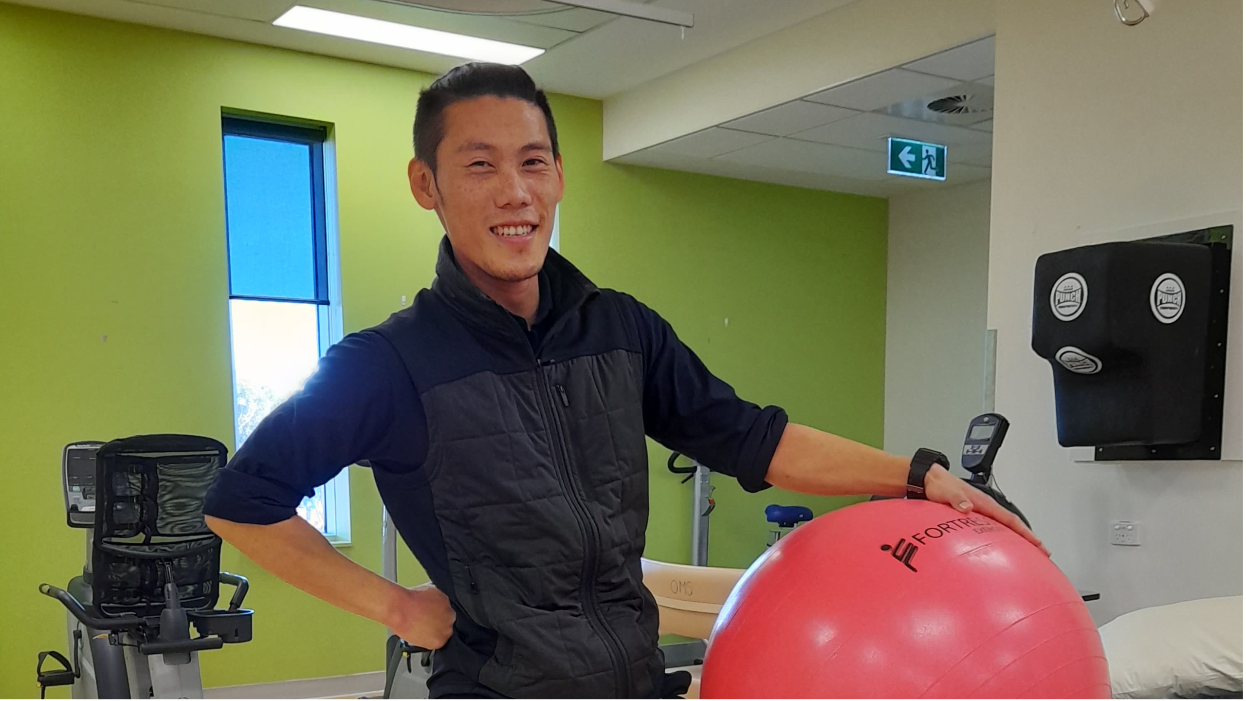 Paul Truong, one of our physiotherapists, is in our rehabilitation gym holding an exercise ball and smiling to the camera.