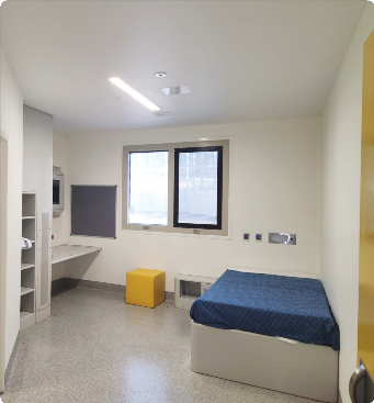 A single bedroom at Dhulwa, with a single bed and desk area as well as some small bookshelves.