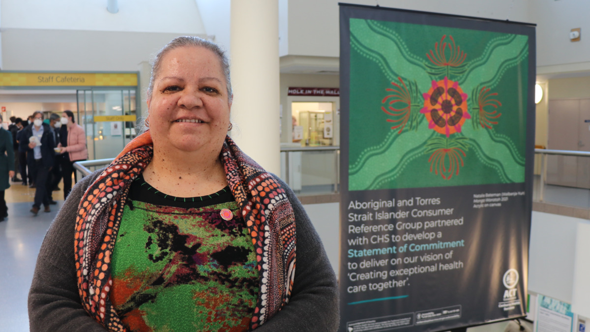 A woman stands in the foreground, smiling, In the background is a large banner showing CHS's Statement of Commitment. It has a beautiful artwork with a Waratah flower on it